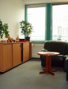 Blick in den Empfangsraum im BZSL e. V. Im Hintergrund befinden sich Fenster. Davor steht links ein Schrank mit Grünpflanzen darauf, daneben ein Tisch und ein Sofa.
