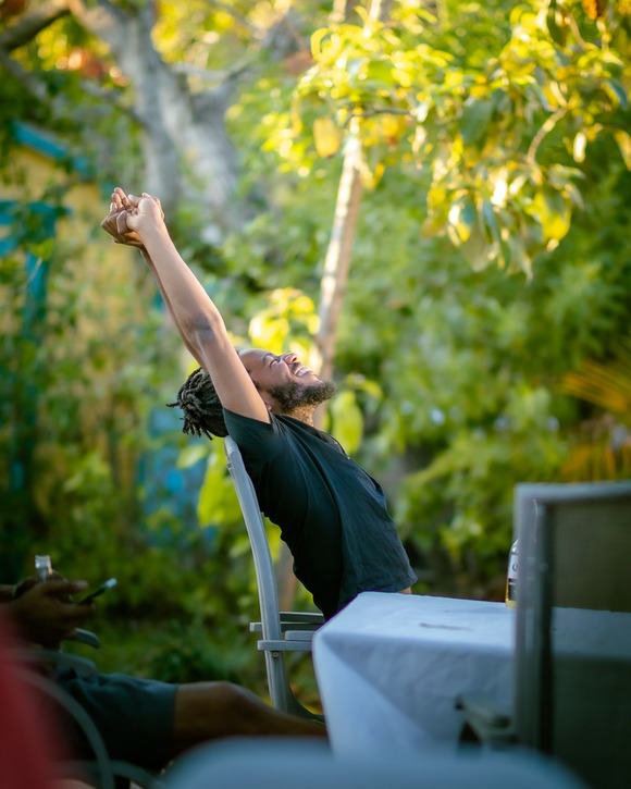 Ein junger Mann sitzt auf einem Stuhl im Garten und streckt entspannt lachend seine Arme nach oben.