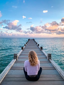 Auf einem Steg, der ins Meer führt, sitzt unter blauem Himmel mit weißen und rosa Wolken eine junge Frau im Schneidersitz und schaut aufs Meer.